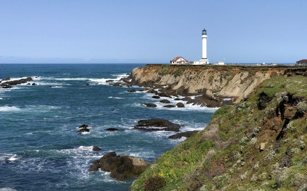 Coastal view featuring a lighthouse on a cliff, overlooking rocky shores and crashing waves under a clear blue sky.