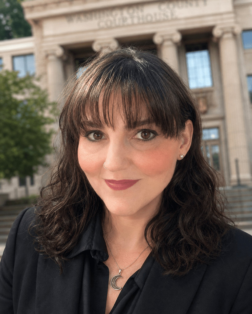Close-up portrait of a woman with dark, wavy hair and a crescent moon necklace, smiling in front of a courthouse building.